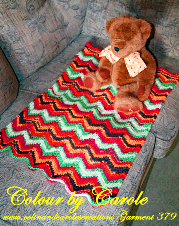 A lovely soft baby blanket hand knitted in bright colours red, orange, green and black. The blanket has a chevron pattern. The blanket measures 45cm by 65cm