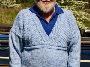 A V neck jumper hand knitted aboard the narrow boat “Emma Maye” in Lancashire by Carole Wareing of Colin and Carole’s Creations. Modelled by Colin, Carole’s husband, it’s a bit tight on him! This garment has been designed and knitted by Carole. It has been created from a shade of “Misty” from J C Brett’s (shade no R5) which is a premium acrylic yarn with a touch of viscose to make it feel nice and soft. The garment is based on a “Scarborough” gansey pattern which has a moss stitch above the yoke on the chest area, while there is a quite distinctive step stitch border between the main body and the arms.